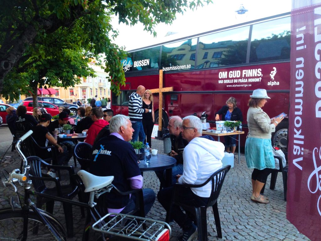 Röd buss i bakgrunden, en samling människor sitter vid sidan om och fikar och samtalar.
